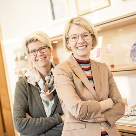 Zwei lächelnde Geschäftsfrauen stehen im modernen Büro vor einem Regal mit Auszeichnungen.
